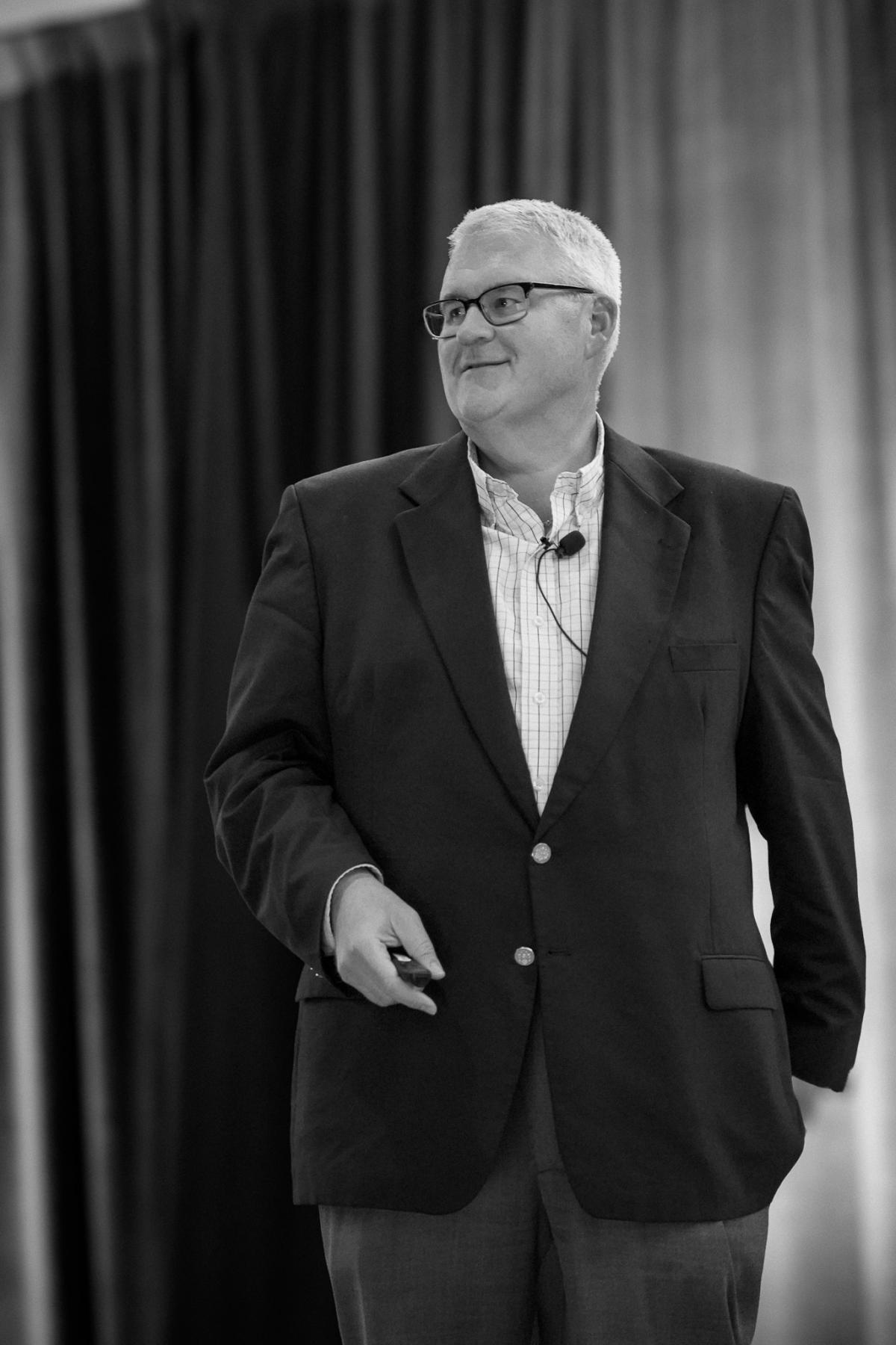 Man in a suit speaking on stage with a microphone headset, smiling.